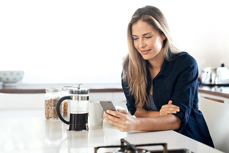 Donna in cucina moderna consulta smartphone accanto a caffettiera, simboleggiando il monitoraggio consumi e risparmio energetico domestico
