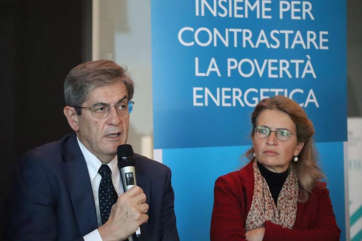 Convegno sulla povertà energetica Italia: uomo con microfono e donna in primo piano davanti a banner blu